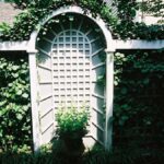 Handcrafted white garden trellis with arched structure and lattice panels supporting climbing plants in an outdoor garden setting.
